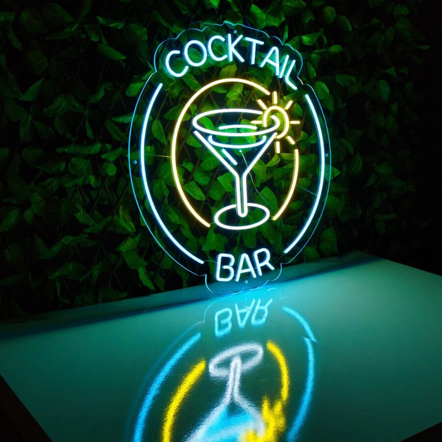 Letrero de Neón Cocktail Bar de una copa de cóctel con las palabras "cocktail bar" sobre un fondo de hojas verdes, con un reflejo en una superficie debajo.
