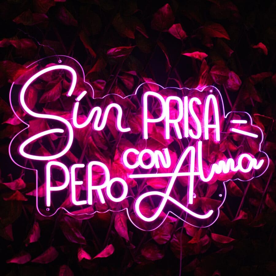 Letrero de Neón Brilla sin miedo con las palabras "sin prisa = pero con alma" en escritura rosa, montado sobre un fondo de hojas oscuras.