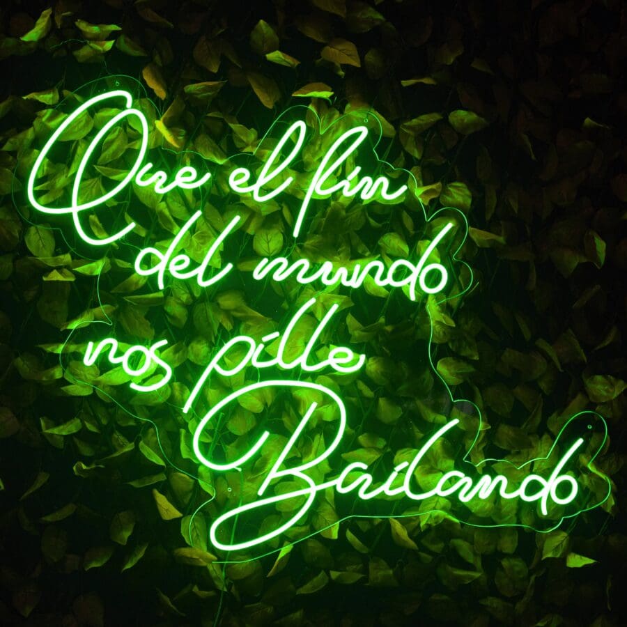 Un Neón Que el fin del mundo NOS pille bailando en texto cursivo en español que dice "Que el fin del mundo nos pille bailando", iluminado en verde brillante sobre un fondo de exuberantes hojas verdes.