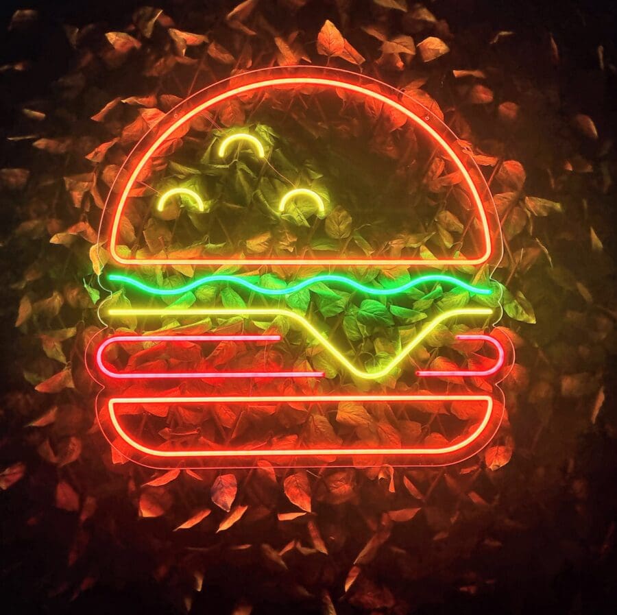 Letrero de neón de una Hamburguesa Neón con lechuga, queso y una hamburguesa, brillando en rojo, amarillo y verde sobre un fondo de hojas.