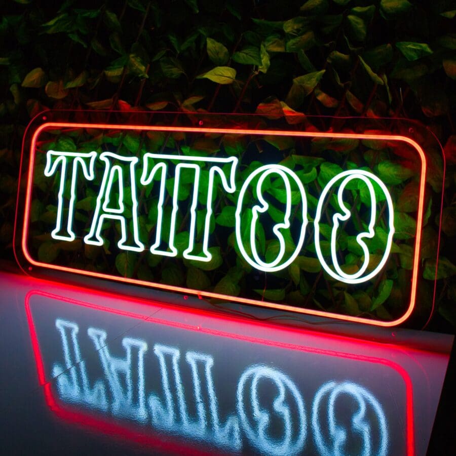 Un letrero de Neón Tattoo brillantemente iluminado que dice "TATTOO" en letras verdes con un borde rojo, colocado contra un fondo de hojas y reflejado en una superficie brillante debajo.