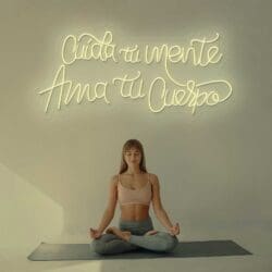 Una mujer haciendo yoga frente al cartel Neón Cuida tu mente, Ama tu cuerpo.
