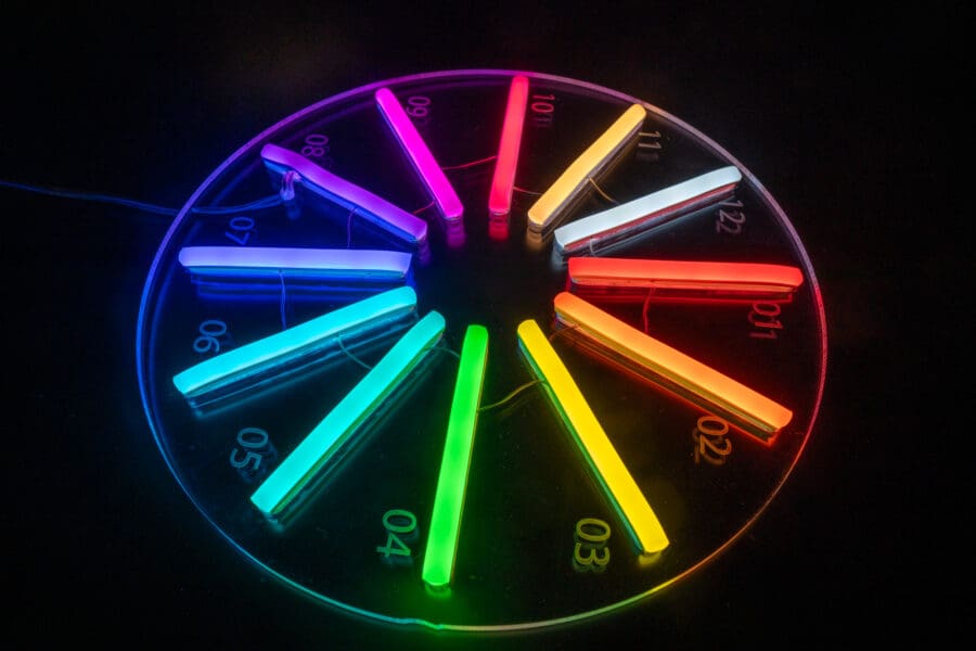 Un reloj LED circular que muestra luces de varios colores que representan cada hora sobre un fondo negro. Los números del 01 al 12 están marcados en la esfera del reloj, creando un efecto de Letras en Neón Personalizadas.