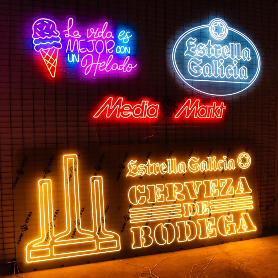 Neón personalizado con texto y logotipos en español en una pared de ladrillos, promocionando la cerveza Estrella Galicia, Media Markt y la frase "la vida es mejor con un helado".