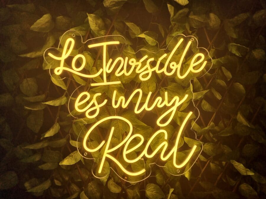 Un letrero de neón amarillo dice "Lo Imposible es muy Real" sobre un fondo de hojas verdes, evocando el misterioso encanto de Neón Lo invisible es muy Real.
