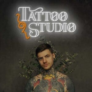 Un hombre con tatuajes parado frente a un letrero de Neón Estudio de tatuaje en un estudio de tatuajes.