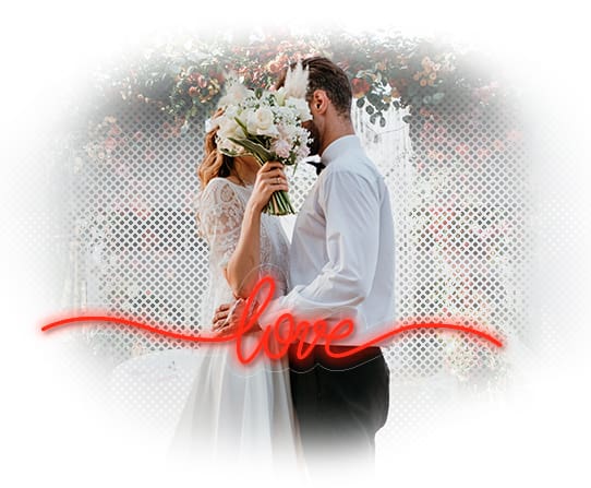Una pareja vestida de novia compartiendo un momento íntimo con un letrero de neón que dice "amor" en primer plano.