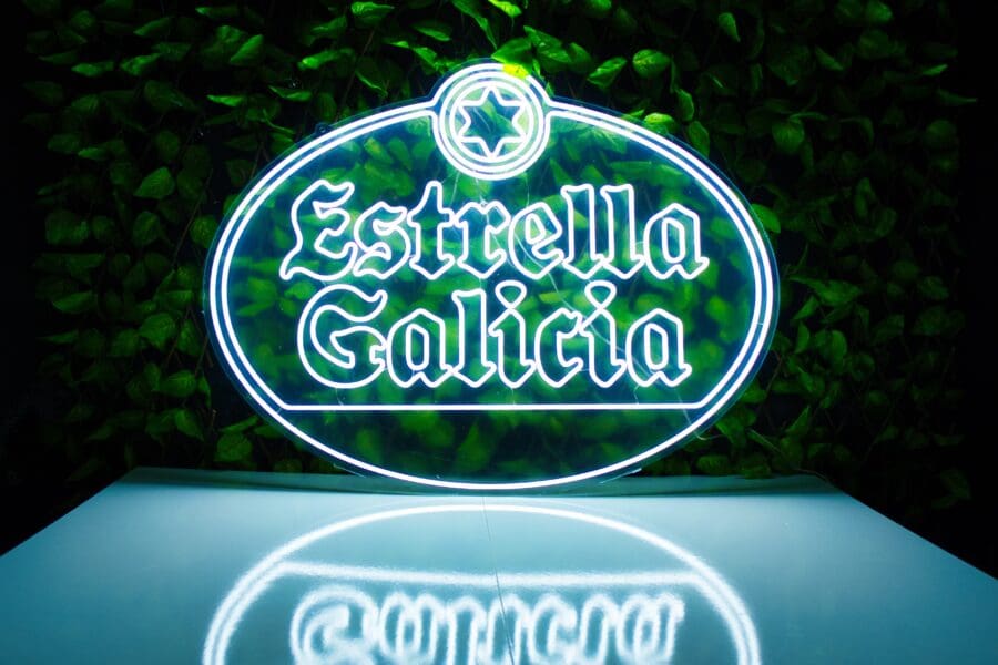Letrero de neón del logo de la cerveza estrella galicia iluminado sobre un fondo de hojas verdes.
