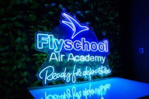 Letrero de neón de la "academia aérea flyschool" con un símbolo de avión sobre un fondo de hojas verdes.