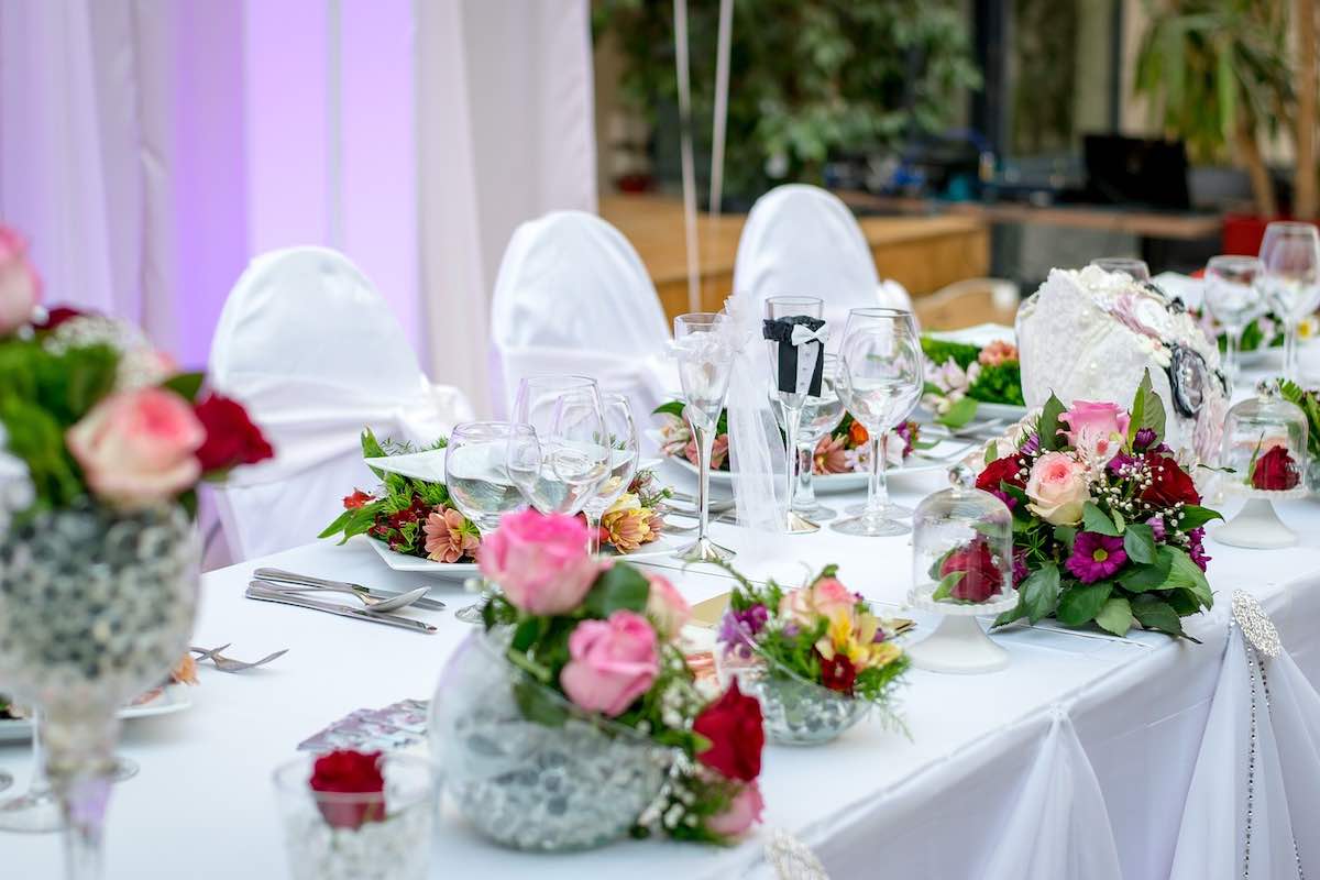 Elegante mesa de recepción nupcial con centros de mesa florales, sillas blancas y cristalería en un lugar con iluminación violeta.