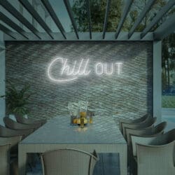 Comedor al aire libre con mesa larga y sillas de mimbre. Un letrero de neón en una pared texturizada dice "Neón Chill Out" con una planta cerca, invitando a los huéspedes a relajarse. ¡No olvides tomarte una selfie en este encantador entorno!