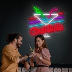 Dos personas chocan copas de cóctel bajo un vibrante letrero de "Neón Cocktails", que presenta un diseño de copa de martini brillante: el momento perfecto para tomarse una selfie.