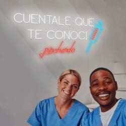 Dos sonrientes trabajadores de la salud vestidos con batas azules se toman una selfie bajo un letrero de neón en español que dice "Neón Cuéntale que te conocí pinchando", junto a la imagen de una jeringa.
