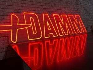 Un letrero de neón rojo en una pared de ladrillos dice "DAMM" con un símbolo de cruz a la izquierda, reflejándose en una superficie brillante debajo.
