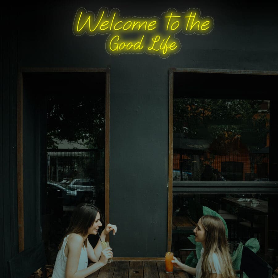 Dos mujeres sentadas en una mesa al aire libre de un restaurante con un letrero de neón Bienvenido a la Buena Vida en lo alto. Uno sostiene una bebida mientras conversan. El vibrante brillo de neón crea el telón de fondo perfecto cuando deciden tomarse una selfie para capturar el momento.