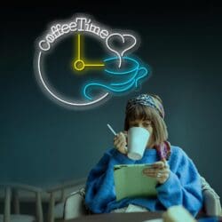 Una persona con un suéter azul y un pañuelo colorido en la cabeza bebe de una taza mientras mira una tableta. Detrás de ellos, un letrero de neón dice "Neón Coffe Time" con un diseño de reloj y una taza de café humeante, creando el escenario perfecto para tomarse una selfie.