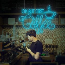 Un barista con camisa negra prepara café con una máquina de café expreso. Sobre él, un letrero de neón dice "Neón OK But First Coffee" con el ícono de una taza de café. Perfecto para esos momentos de Instagram, ¡no olvides tomarte una selfie!