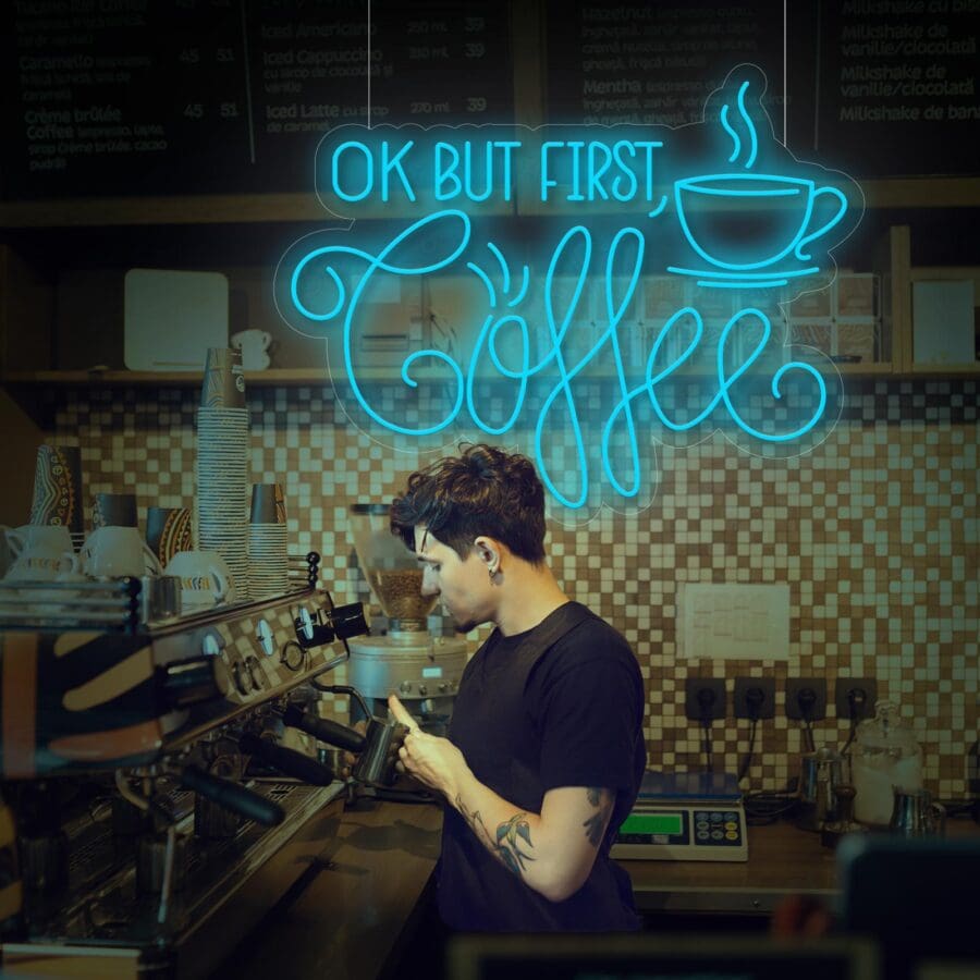 Un barista con camisa negra prepara café con una máquina de café expreso. Sobre él, un letrero de neón dice "Neón OK But First Coffee" con el ícono de una taza de café. Perfecto para esos momentos de Instagram, ¡no olvides tomarte una selfie!
