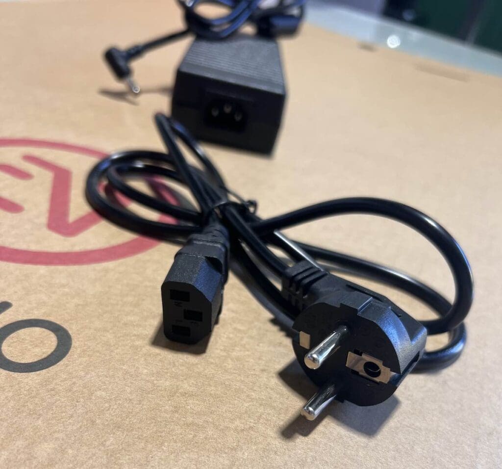 Sobre una superficie de cartón se coloca un cable de alimentación eléctrica negro con conector IEC 60320 C13 y enchufe europeo, con un adaptador de corriente negro al fondo.