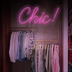 Un letrero de neón que dice "¡Chic!" cuelga sobre un perchero con varias camisas y blusas en colores pastel y claros. El fondo presenta una pared con texturas discretas.