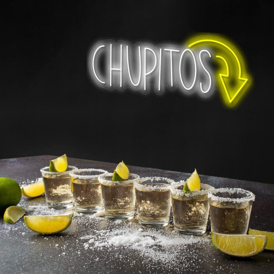 Cinco vasos de chupito con bordes de sal y gajos de lima están alineados sobre una mesa con sal y rodajas de lima esparcidas, debajo de un letrero de neón que dice "CHUPITOS" con una flecha apuntando hacia abajo.