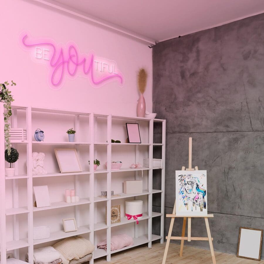 Una habitación con estantes blancos que contienen diversos artículos, un caballete con una pintura colorida y un letrero de neón en la pared que dice "BEYOUTIFUL".