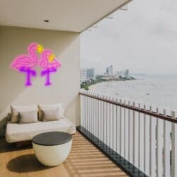 Un balcón junto a la playa con un sofá, una mesa redonda y un letrero de neón de dos flamencos en la pared ofrece vistas al horizonte de la ciudad y al océano.