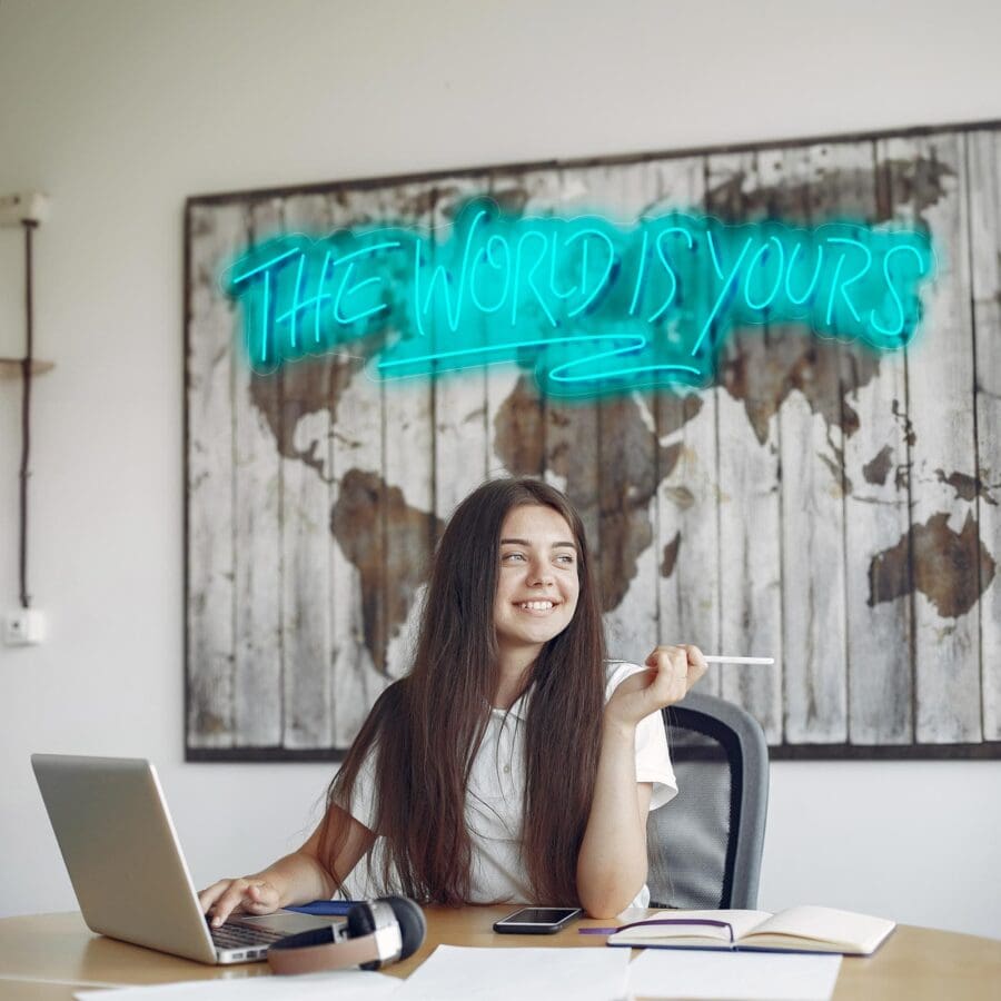 Una persona está sentada en un escritorio trabajando con un ordenador portátil, con un bolígrafo en la mano y cuadernos y auriculares cerca. De fondo se ven un mapa del mundo y un cartel de neón que dice "El mundo es tuyo".