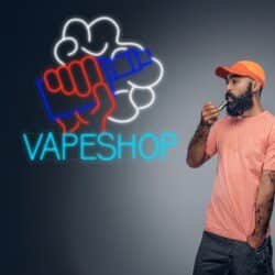 Hombre con una camisa rosa y una gorra naranja vapeando junto a un letrero de neón que dice "VAPESHOP" con una imagen de un puño sosteniendo un vaporizador.