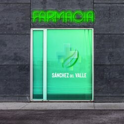 Letrero de neón que dice "Farmacia" sobre una puerta de vidrio verde con una cruz médica y "Sánchez del Valle" impreso en ella. La puerta está empotrada en una pared de hormigón gris.