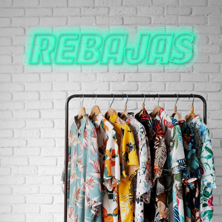 Sobre una pared de ladrillos blancos se exhibe un perchero con varias camisetas de colores y estampados florales. Encima, un letrero de neón dice "REBAJAS".