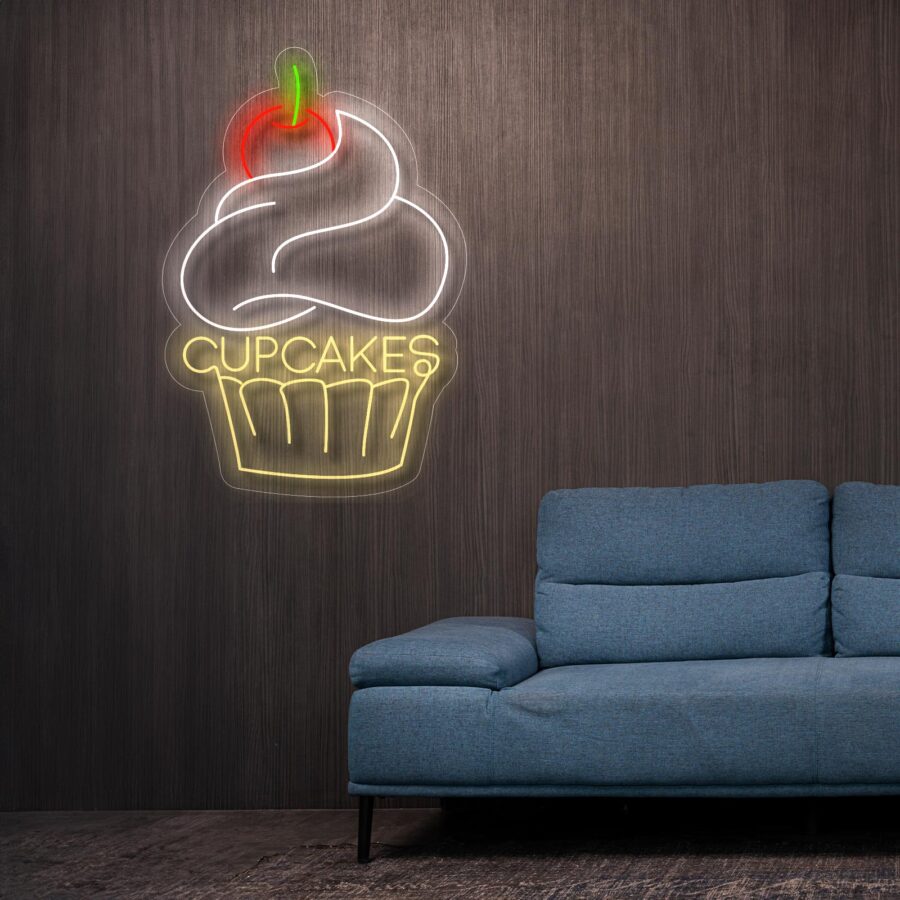 Un letrero de neón de un cupcake con la palabra "Cupcakes" en luz blanca y amarilla cuelga en una pared de madera oscura sobre un sofá azul.