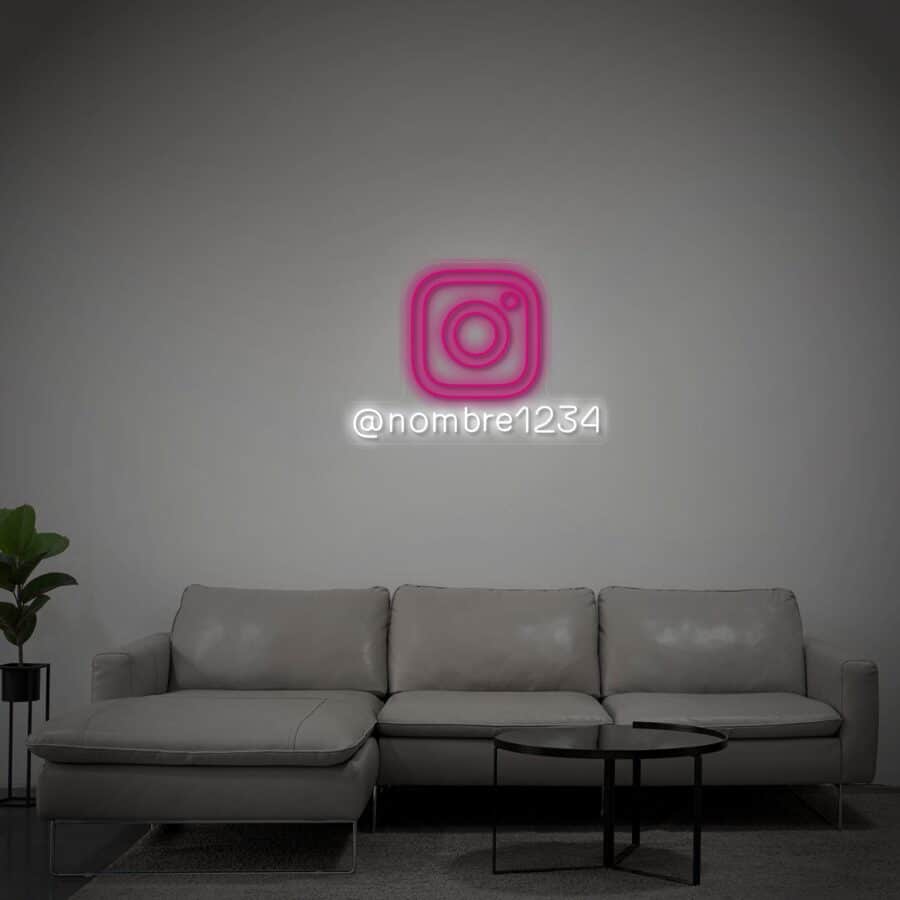 Un sofá gris se apoya contra una pared lisa con un logotipo de Instagram en neón rosa y el nombre de usuario "@nombre1234" encima. Hay una pequeña planta a la izquierda y mesas redondas frente al sofá.