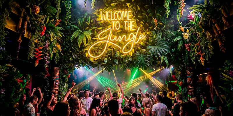 Discoteca abarrotada con decoración de temática selvática, letrero de neón "Welcome to the Jungle", vegetación en lo alto y luces de colores que iluminan a la gente que baila.