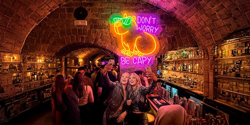 Dos mujeres se toman un selfie en un bar abarrotado con arcos de ladrillo y un letrero de neón con un carpincho que dice: "No te preocupes, sé carpincho".