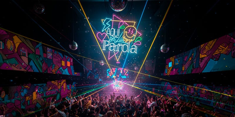 Una gran multitud baila en un club nocturno con coloridos carteles de neón, vibrantes obras de arte en las paredes, bolas de discoteca y luces láser que iluminan la escena.
