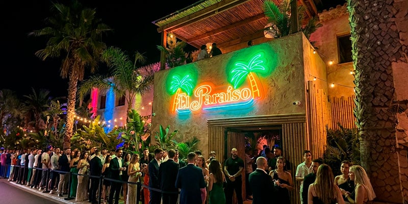Una multitud hace cola ante una discoteca de temática tropical llamada "El Paraíso" por la noche, con luces de neón y palmeras a la vista.