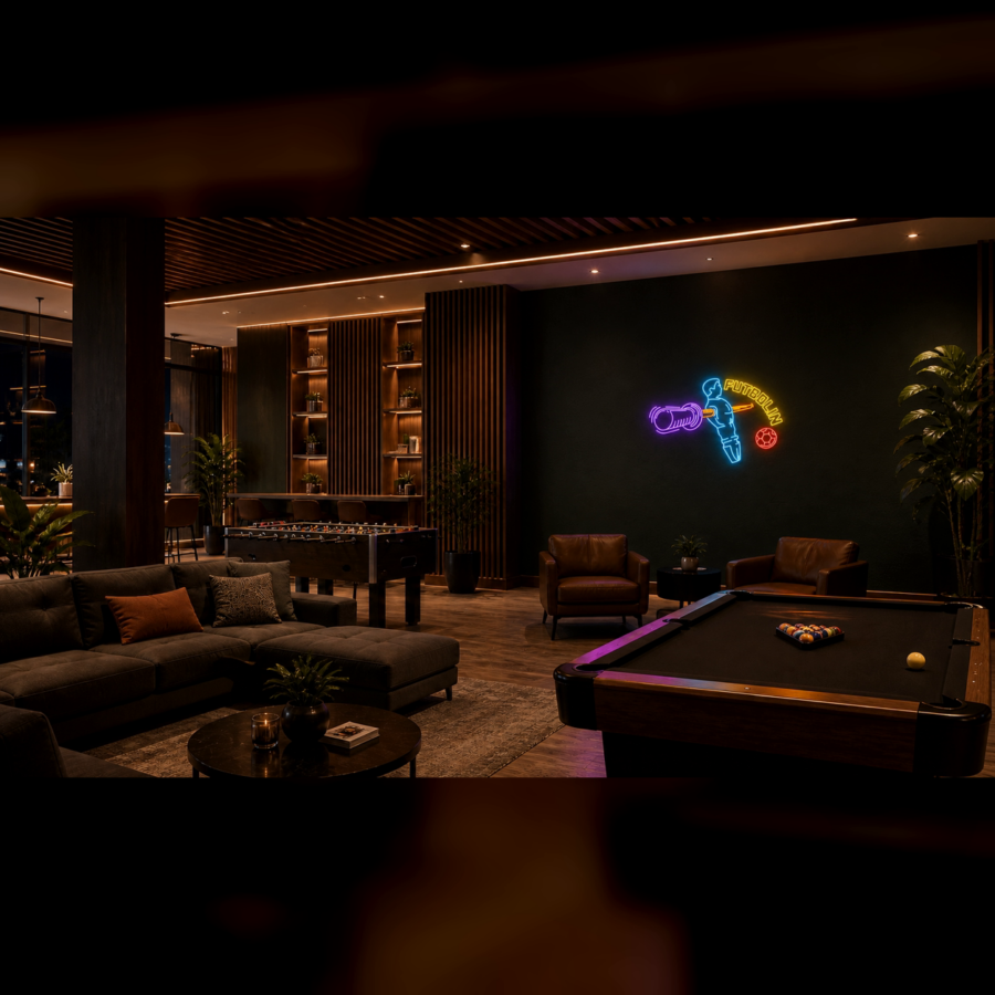 Un salón moderno cuenta con una mesa de billar, un futbolín, sofás marrones, sillones y un cuadro de neón en una pared oscura, con una iluminación cálida y plantas de interior.