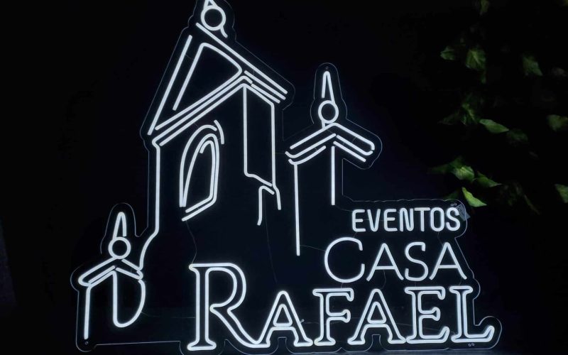 Letrero de neón que representa un edificio con el texto "EVENTOS CASA RAFAEL" iluminado sobre un fondo oscuro.