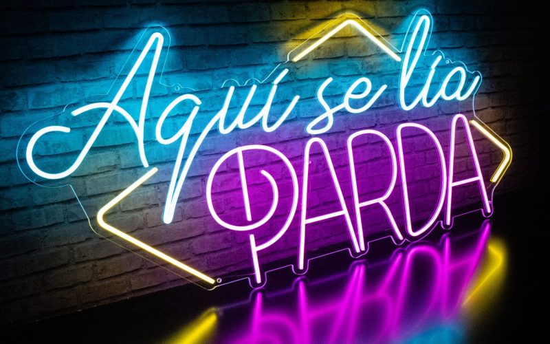 Alquiler Neón Aquí se Lía Parda con el texto en español "Aquí se lía parda", iluminado en azul, amarillo y rosa sobre un fondo de pared de ladrillos.