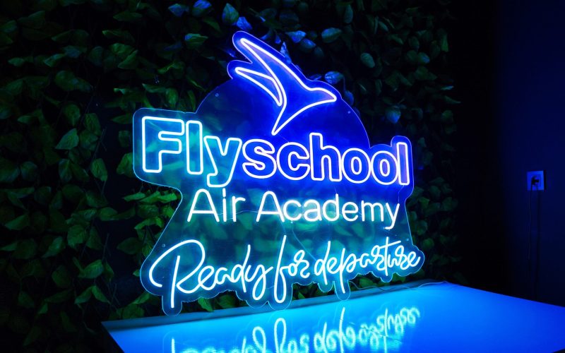 Letrero de neón de la "academia aérea flyschool" con un símbolo de avión sobre un fondo de hojas verdes.